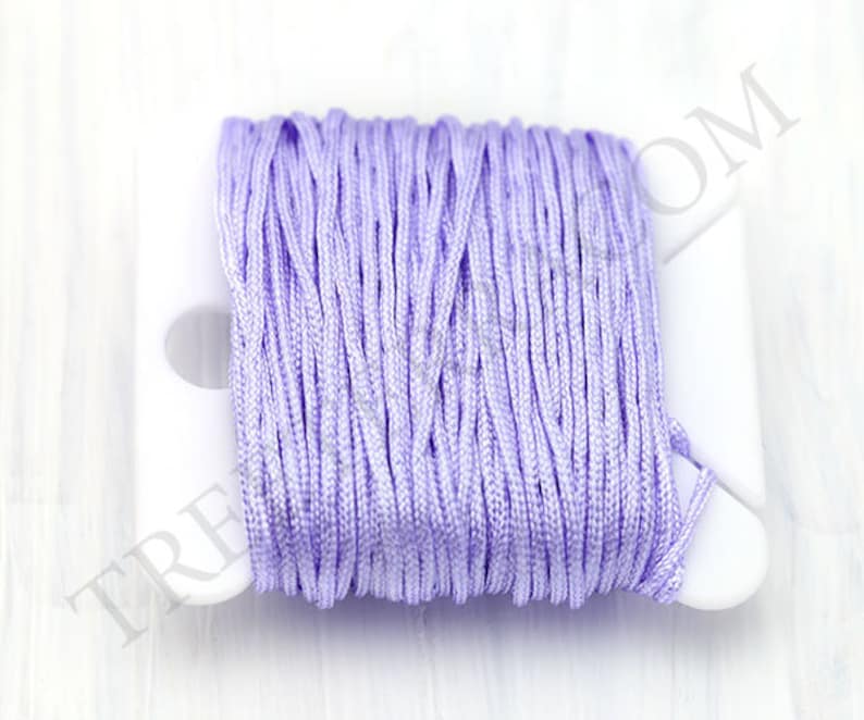 Puede incluir: Un carrete de cord&oacute;n de sat&eacute;n de color lavanda. El cord&oacute;n est&aacute; enrollado firmemente en un carrete de pl&aacute;stico blanco. La textura del cord&oacute;n es suave y parece estar hecho de muchos hilos finos. El carrete est&aacute; sobre un fondo blanco.