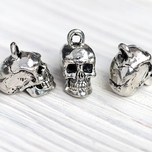 May include: Three silver-colored skull charms with a loop at the top for hanging. The skulls are detailed with eye sockets, nasal cavity, and teeth. The charms are displayed on a white, textured surface.