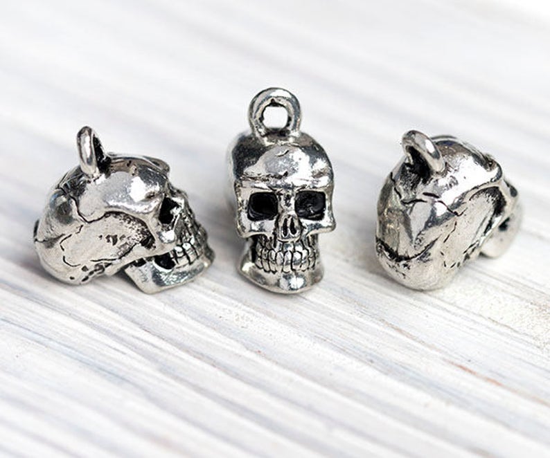May include: Three silver-colored skull charms with a loop at the top for hanging. The skulls are detailed with eye sockets, nasal cavity, and teeth. The charms are displayed on a white, textured surface.