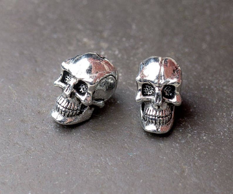 Puede incluir: Dos peque&ntilde;as cuentas con forma de calavera de color plateado. Cada calavera tiene detalles, incluyendo cuencas de los ojos, una cavidad nasal y una dentadura completa. Las calaveras probablemente se utilizan para joyer&iacute;a o proyectos de artesan&iacute;a.