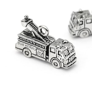 May include: Silver-colored miniature fire truck charm with the text "OUR HEROES" on the side. The truck features a ladder and the letters "F.D." on the front. The charm is detailed and has a vintage appearance.