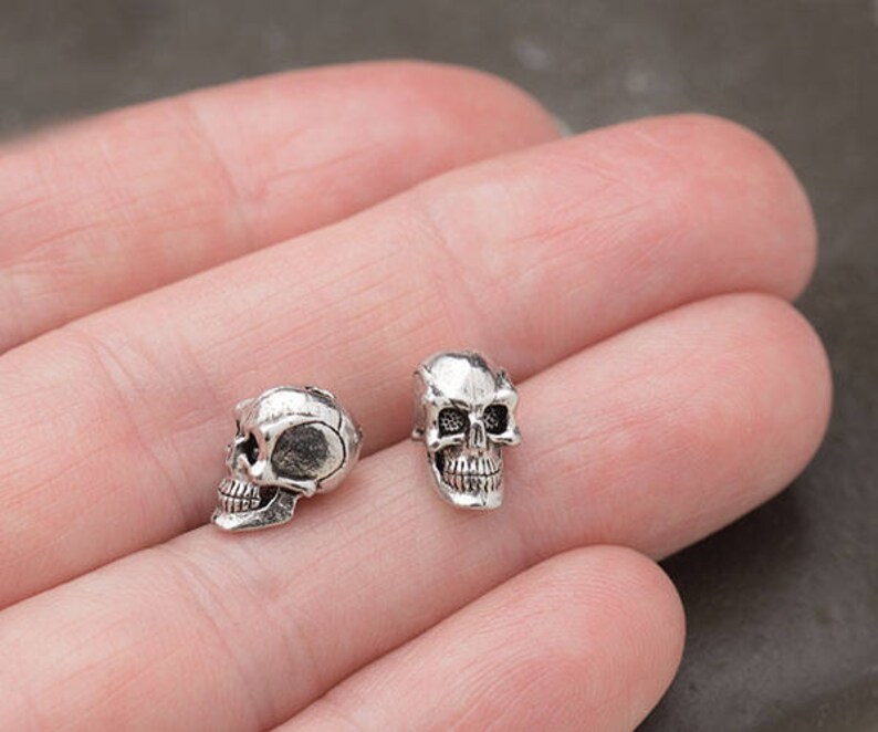 Puede incluir: Dos peque&ntilde;as cuentas de calavera de color plateado. Las calaveras tienen detalles, incluyendo cuencas de los ojos, una cavidad nasal y dientes. Estas cuentas son probablemente para la fabricaci&oacute;n de joyas u otros proyectos de artesan&iacute;a.