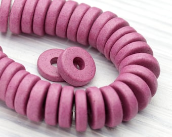 Puede incluir: Primer plano de un collar de cuentas moradas en forma de rosquilla. Dos cuentas sueltas están al lado del collar. Las cuentas son de color sólido y tienen un acabado mate. El fondo es de vetas de madera clara.