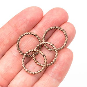 Pode incluir: Close-up de vários anéis de metal torcido de cor bronze. Os anéis são circulares e têm uma textura semelhante à de uma corda. Os anéis são segurados na mão de uma pessoa contra um fundo branco.
