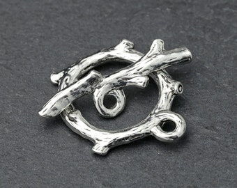 May include: A silver-toned toggle clasp with a rustic, twig-like design. The clasp features a circular loop and a bar, both textured to resemble tree branches. The clasp is designed for jewelry making and crafting.