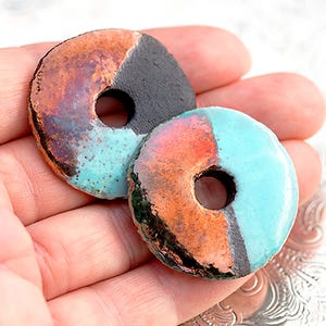 Puede incluir: Dos cuentas de cerámica en forma de rosquilla con un agujero en el centro. Una cuenta está dividida en secciones negras y naranjas, la otra en turquesa y naranja. Las cuentas tienen un acabado texturizado y vidriado, y se sostienen en una mano.
