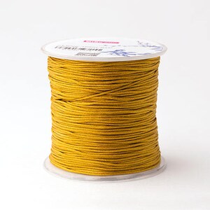 Puede incluir: Un carrete de cord&oacute;n amarillo dorado, bien enrollado y exhibido sobre un fondo blanco. El cord&oacute;n parece liso y est&aacute; enrollado en una bobina de pl&aacute;stico transparente. La parte superior del carrete tiene detalles impresos.
