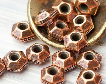 May include: Close-up of copper-colored, geometric beads with a hexagonal shape and a central hole. The beads have a textured surface and are arranged on a small, gold-toned scoop and a white surface. These beads are ideal for jewelry making.