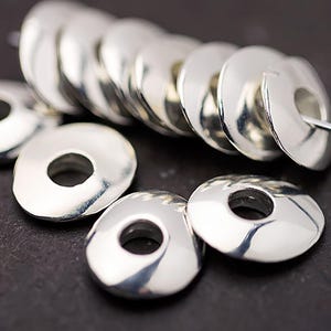 May include: Close-up of several shiny, silver-colored metal disc beads with a hole in the center. The beads are arranged in a row and scattered, reflecting light. These beads are suitable for jewelry making.