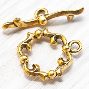 May include: Ornate gold-coloured toggle clasp jewellery findings. The clasp features a circular design with decorative flourishes and a matching bar. The metal has an antique finish, suggesting a vintage or handcrafted aesthetic. Ideal for jewellery making.