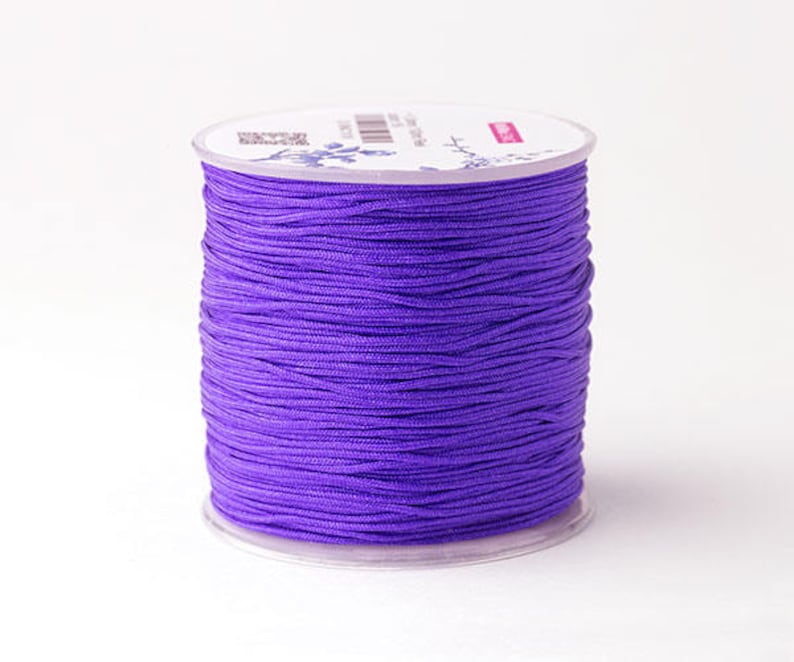 Puede incluir: Un carrete de cord&oacute;n morado vibrante, bien enrollado y exhibido sobre una base de pl&aacute;stico transparente. El cord&oacute;n parece liso y es adecuado para varios proyectos de manualidades. El carrete se encuentra sobre un fondo blanco liso.
