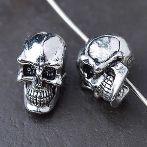 Puede incluir: Dos cuentas plateadas con forma de calavera con detalles faciales, incluyendo cuencas de los ojos, cavidad nasal y dentadura completa. Las cuentas est&aacute;n ensartadas en un fino alambre plateado, lo que sugiere su uso en joyer&iacute;a o manualidades.