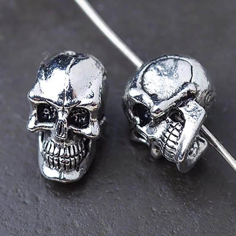 Puede incluir: Dos cuentas plateadas con forma de calavera con detalles faciales, incluyendo cuencas de los ojos, cavidad nasal y dentadura completa. Las cuentas est&aacute;n ensartadas en un fino alambre plateado, lo que sugiere su uso en joyer&iacute;a o manualidades.