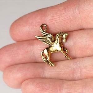 Op de afbeelding: Een goudkleurige Pegasus-bedel met uitgespreide vleugels. Het mythische gevleugelde paard is gedetailleerd met een lus aan de bovenkant om aan een ketting of armband te bevestigen. De bedel wordt in een hand gehouden.