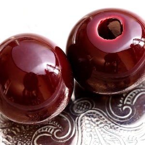 Könnte beinhalten: Zwei glänzende, dunkelrote Kugeln mit einem Loch in einer. Die Kugeln liegen auf einer dekorativen, silberfarbenen Oberfläche. Die Objekte reflektieren das Licht und erzeugen Glanzlichter und Schatten.