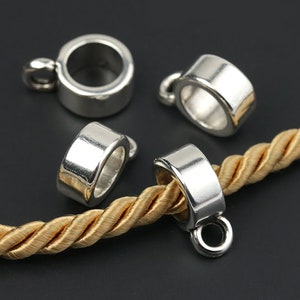 Puede incluir: Cuatro hallazgos de joyería de metal plateado, cada uno con un lazo para sujetar a una cadena o cordón. Un hallazgo está unido a un cordón trenzado de color dorado. Los hallazgos son cilíndricos con un acabado liso y brillante.