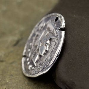 May include: A silver-colored, oval-shaped coin with an embossed design featuring a profile of a person. The coin has a hole at the top and a textured surface, suggesting an antique or vintage aesthetic. The coin is slightly angled, revealing its thickness.
