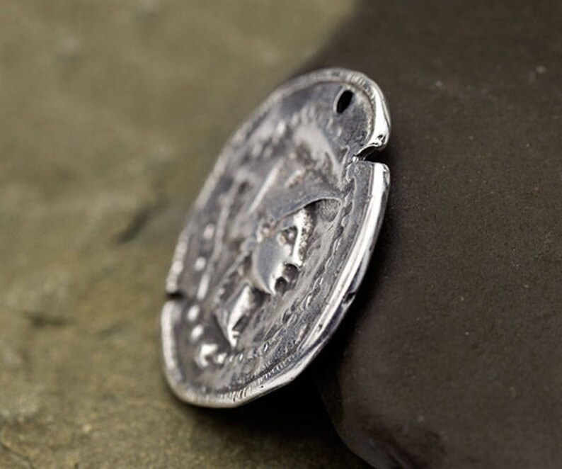 May include: A silver-colored, oval-shaped coin with an embossed design featuring a profile of a person. The coin has a hole at the top and a textured surface, suggesting an antique or vintage aesthetic. The coin is slightly angled, revealing its thickness.
