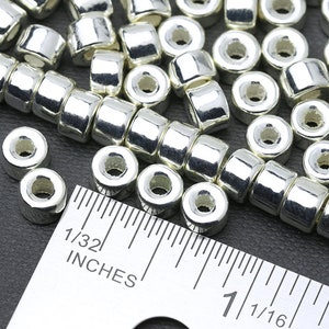 May include: Close-up of shiny silver-colored metal beads. The beads are cylindrical with a hole running through the center. A ruler is placed next to the beads, with measurements in inches. The beads are likely for jewelry making or crafts.