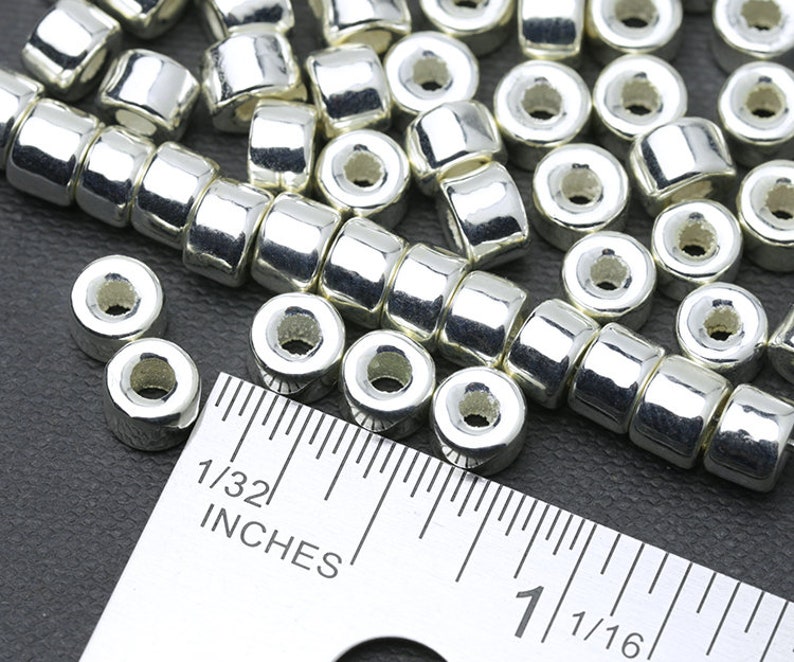 May include: Close-up of shiny silver-colored metal beads. The beads are cylindrical with a hole running through the center. A ruler is placed next to the beads, with measurements in inches. The beads are likely for jewelry making or crafts.
