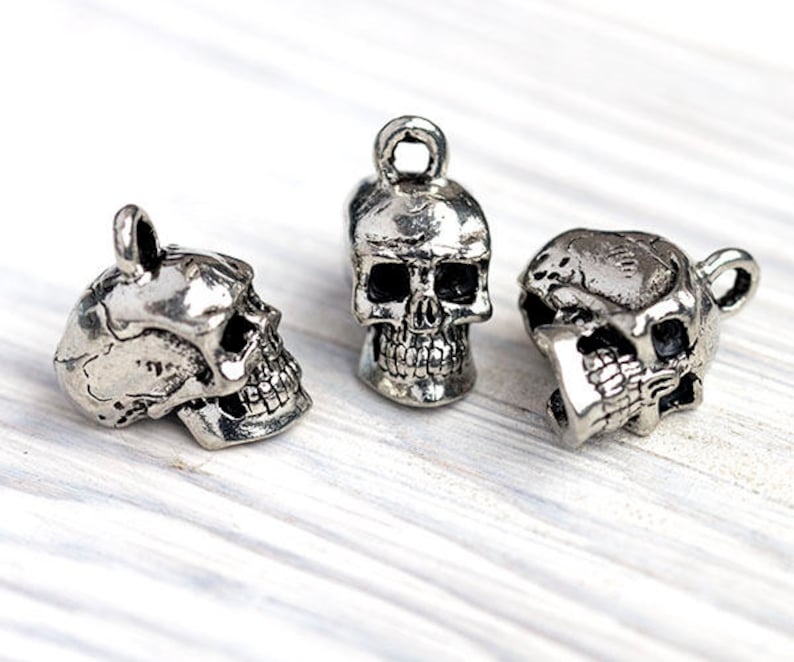 May include: Three silver-colored skull charms with a loop at the top for hanging. The skulls have detailed facial features, including eye sockets, a nose cavity, and teeth. The charms are displayed on a white wooden surface.