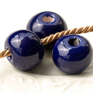 May include: Three glossy, navy blue ceramic beads with a hole through the centre, strung on a twisted gold-coloured cord. The beads have a smooth, reflective surface, against a white background.