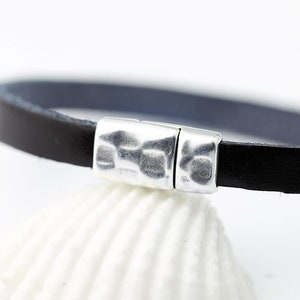 Puede incluir: Una pulsera de cuero negro con un cierre magn&eacute;tico plateado. La pulsera es delgada y tiene un interior azul oscuro. El cierre tiene una textura martillada. La pulsera se muestra sobre una superficie blanca.