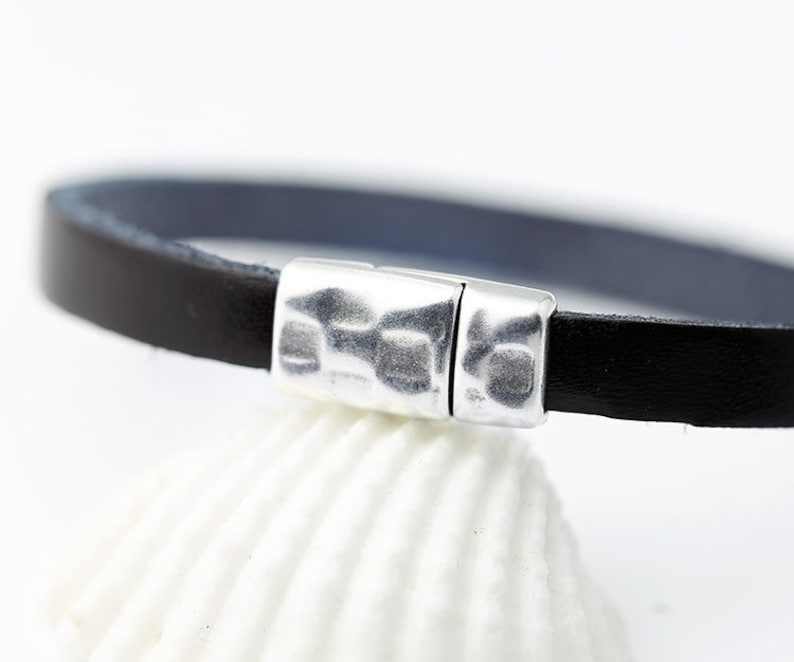 Puede incluir: Una pulsera de cuero negro con un cierre magn&eacute;tico plateado. La pulsera es delgada y tiene un interior azul oscuro. El cierre tiene una textura martillada. La pulsera se muestra sobre una superficie blanca.