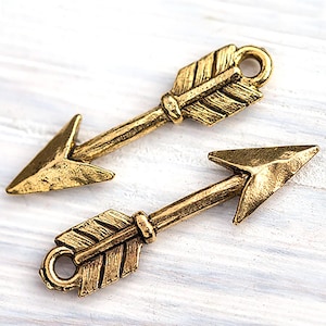May include: Two antique gold-coloured arrow charms. Each arrow features a pointed arrowhead, a feathered fletching, and a loop for attaching to jewellery or crafts. The arrows have a textured, aged appearance.