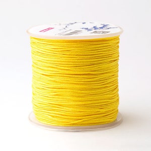 Puede incluir: Una bobina de hilo amarillo brillante. El hilo est&aacute; enrollado firmemente alrededor de un n&uacute;cleo de cart&oacute;n blanco con una base de pl&aacute;stico transparente. El hilo parece liso y tiene un ligero brillo. La bobina est&aacute; sobre un fondo blanco liso.
