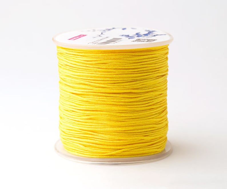 Puede incluir: Una bobina de hilo amarillo brillante. El hilo est&aacute; enrollado firmemente alrededor de un n&uacute;cleo de cart&oacute;n blanco con una base de pl&aacute;stico transparente. El hilo parece liso y tiene un ligero brillo. La bobina est&aacute; sobre un fondo blanco liso.