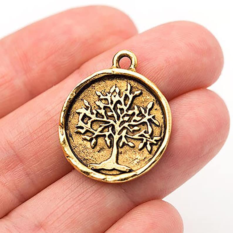 May include: A round, antique gold-colored charm with a tree design. The tree is in the center of the charm, with detailed branches and leaves. The charm has a loop at the top for attaching to a necklace or bracelet. The charm is approximately 1 inch in diameter.
