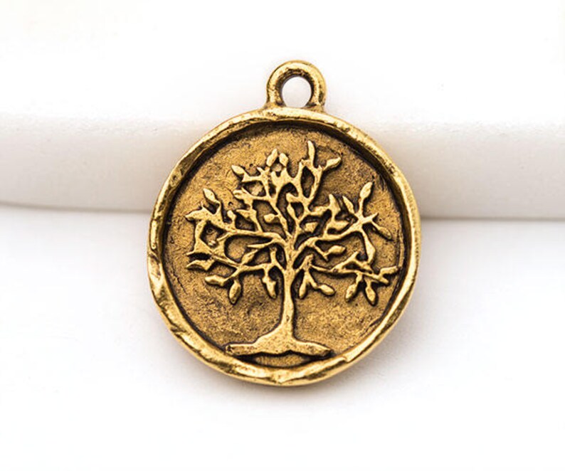 May include: A round, antique gold-colored charm featuring a detailed tree design. The tree has a prominent trunk and branches with leaves, set within a circular frame. The charm has a loop at the top for attaching to jewelry.