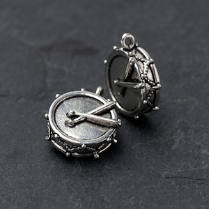 Può includere: Due ciondoli a forma di tamburo color argento. Un ciondolo presenta le bacchette incrociate sulla pelle del tamburo. L'altro ciondolo mostra il retro del tamburo con un disegno a forma di stella. Entrambi i ciondoli hanno un anello per attaccare a una collana o un braccialetto.