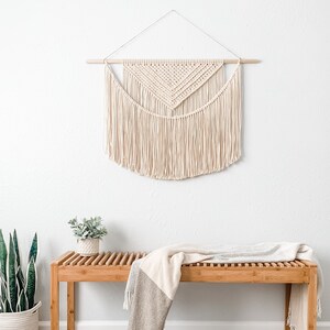 May include: A white macrame wall hanging with a geometric design hangs above a wooden bench with a gray and white throw blanket draped over it. A potted plant sits on the floor next to the bench.