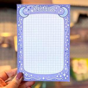 May include: A purple notepad with a grid pattern and the word "Notes" at the top. The notepad is decorated with crescent moons and stars. The notepad is held by a person.