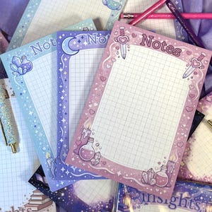 May include: A collection of decorative notepads with grid paper. Each notepad features a different pastel color border with fantasy-themed illustrations, including potion bottles, crystals, and a crescent moon. The word "Notes" is printed on the top of one notepad.