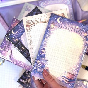May include: A collection of purple and blue notepads with the word "insights" printed on them. Each notepad features illustrations of books, moons, stars, and lanterns. The notepads have a grid pattern and a cup of coffee on one of them.