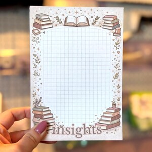 May include: A rectangular notepad with a grid pattern and the word "insights" at the bottom. The notepad is decorated with illustrations of stacked books, open books, and floral elements in shades of brown and beige.