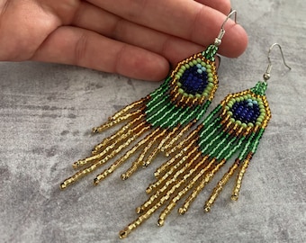 May include: A pair of long, beaded earrings with a peacock eye design. The earrings are made with gold, green, and blue beads and have a silver hook.