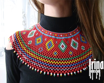 May include: A red, blue, green, yellow, and white beaded necklace with a geometric pattern. The necklace is worn by a person with long brown hair.