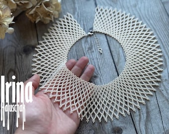 May include: Cream-colored beaded necklace with a geometric design. The necklace is held in a hand, showcasing its intricate pattern and the delicate beads. The necklace has a silver clasp. The name "Irina Haluschak" is written in white on the left side.
