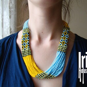 May include: A woman wearing a blue shirt with a multi-strand beaded necklace. The necklace is made of yellow, blue, and black beads and has a geometric pattern. The text "Irina Haluschak" is visible in the lower right corner of the image.