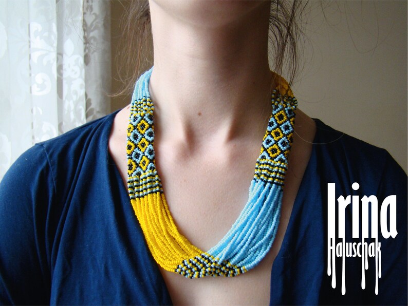 May include: A woman wearing a blue shirt with a multi-strand beaded necklace. The necklace is made of yellow, blue, and black beads and has a geometric pattern. The text "Irina Haluschak" is visible in the lower right corner of the image.