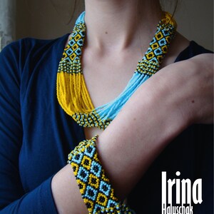 May include: A woman wearing a blue shirt with a long beaded necklace and a matching beaded bracelet. The necklace and bracelet are made of blue, yellow, and black beads. The beads are arranged in a geometric pattern. The woman's name, "Irina Haluschak", is written in white letters on a black background.