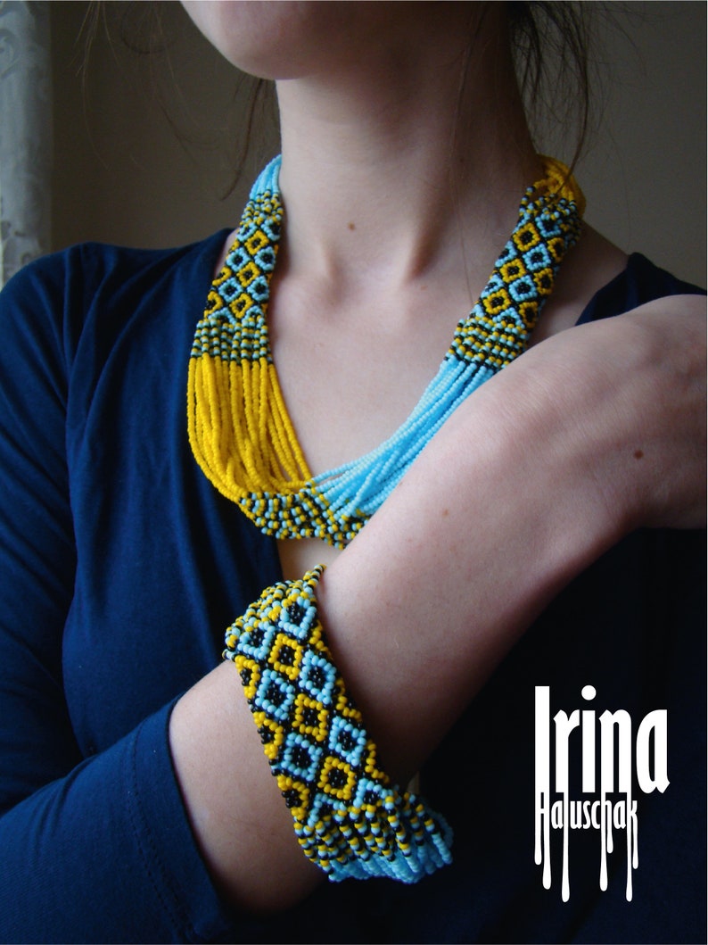 May include: A woman wearing a blue shirt with a long beaded necklace and a matching beaded bracelet. The necklace and bracelet are made of blue, yellow, and black beads. The beads are arranged in a geometric pattern. The woman's name, "Irina Haluschak", is written in white letters on a black background.