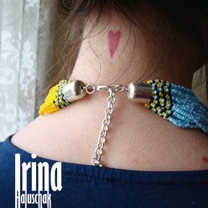 May include: A woman wearing a blue shirt with a beaded necklace featuring yellow, blue, and black beads. The necklace has a silver chain attached to it. The woman has a small red heart drawn on her neck.