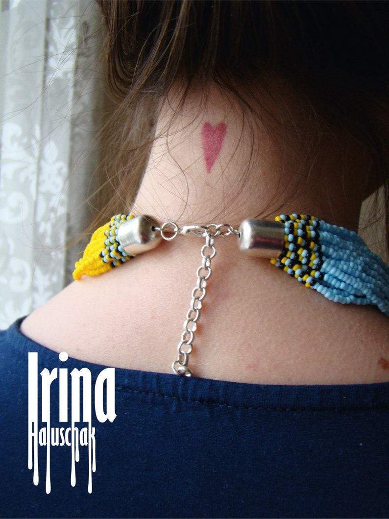 May include: A woman wearing a blue shirt with a beaded necklace featuring yellow, blue, and black beads. The necklace has a silver chain attached to it. The woman has a small red heart drawn on her neck.