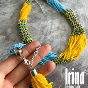 May include: A long beaded necklace with a yellow and blue color scheme. The necklace has a tassel at the end and a silver clasp. The necklace is made of small beads and has a geometric pattern.