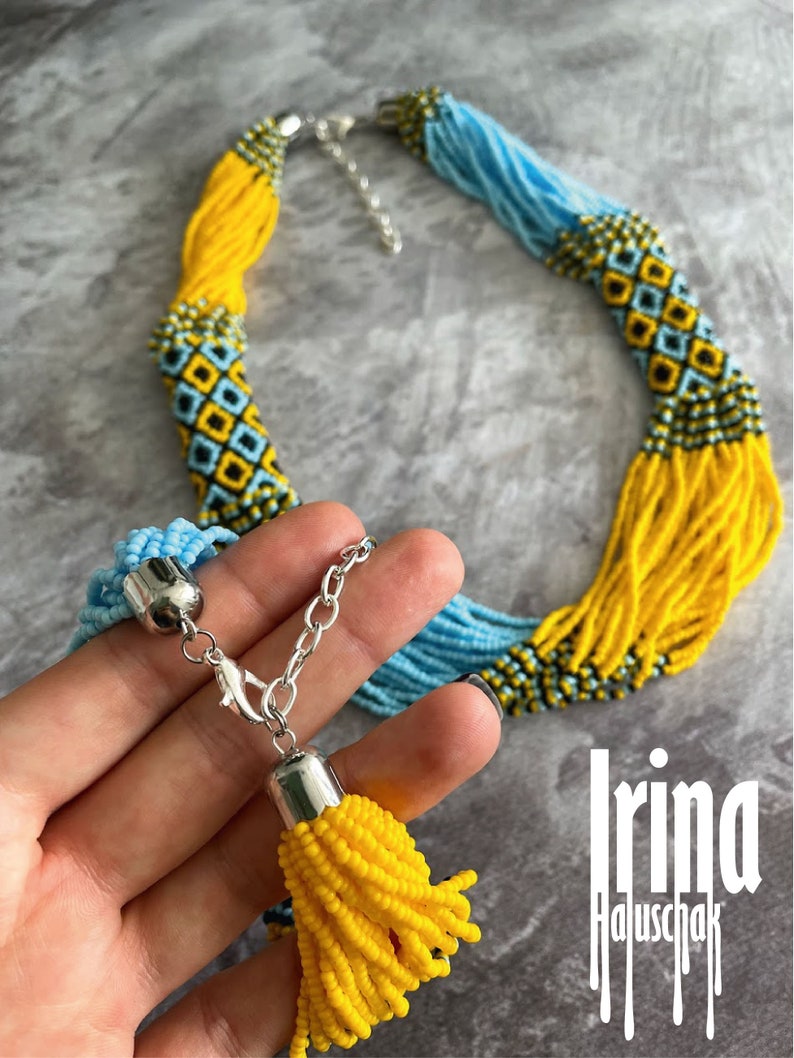 May include: A long beaded necklace with a yellow and blue color scheme. The necklace has a tassel at the end and a silver clasp. The necklace is made of small beads and has a geometric pattern.